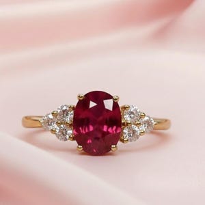 May include: A gold ring with a large oval ruby centre stone, and clusters of smaller white gemstones on either side. The ring has a polished gold band. The background is a soft pink.