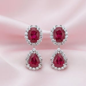 May include: A pair of elegant drop earrings featuring oval and pear-shaped ruby gemstones. Each ruby is surrounded by a halo of small, sparkling diamonds. The earrings are set in a silver metal and hang from a delicate post.