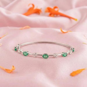 Puede incluir: Una pulsera de plata con esmeraldas ovaladas y piedras redondas transparentes engastadas a lo largo de la banda. La pulsera se exhibe sobre una superficie de satén rosa con pétalos de flores naranjas. Es una joya.