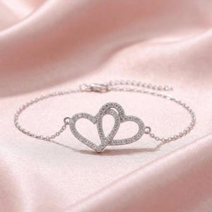 Könnte beinhalten: Ein silbernes Armband mit zwei ineinandergreifenden herzförmigen Anhängern, die jeweils mit kleinen, glitzernden Steinen besetzt sind. Das Armband liegt auf einem weichen, rosa Stoff, was eine romantische und elegante Ästhetik erzeugt.