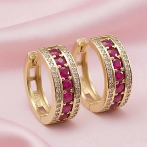 May include: A pair of gold-colored hoop earrings. Each earring features a row of round, deep red gemstones and a row of smaller, clear gemstones. The earrings are displayed on a pink, satin-like surface.