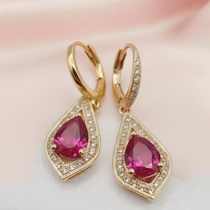 May include: A pair of gold-colored hoop earrings with a teardrop-shaped pendant. Each pendant features a large, vibrant pink gemstone surrounded by small, clear stones. The earrings are designed with a detailed, ornate setting.
