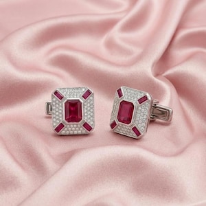 May include: A pair of rectangular cufflinks featuring a central emerald-cut red gemstone surrounded by a border of small white pavé stones and four baguette-cut red gemstones at the corners, set in a polished silver-toned metal frame.