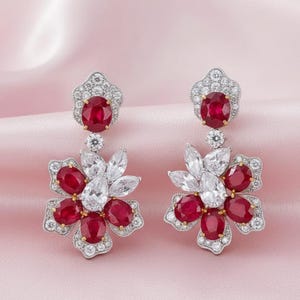 May include: A pair of flower-shaped earrings. Each earring has an oval ruby at the top, a smaller diamond-like stone, and a flower design with ruby and diamond-like stones. The earrings have a silver and gold-tone metal setting.