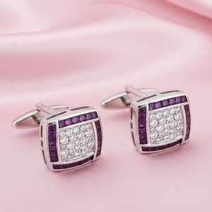 May include: Silver cufflinks with a square design. Each cufflink has a central cluster of small, white stones, framed by a border of rectangular, purple stones. The cufflinks are displayed on a soft pink background.