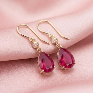 May include: A pair of gold-tone drop earrings featuring pear-shaped, deep red faceted stones suspended from a small, clear round stone. The earrings have curved wire hooks and are crafted with a polished metal finish for a refined look.