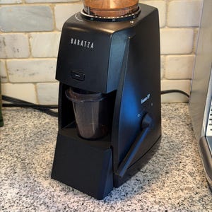 May include: Black Baratza Encore ESP coffee grinder with a clear hopper filled with coffee beans. The grinder has a dial for grind settings and a portafilter holder. The brand name is printed on the front.