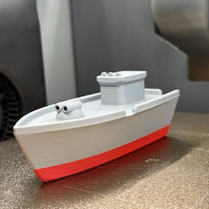 May include: A 3D-printed model of a boat, featuring a gray hull with a red waterline and a white deck. The boat has a small cabin and two cylindrical structures on the bow. A toy or decorative item.