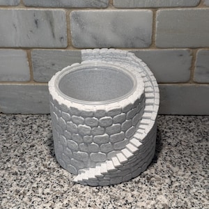 May include: A gray, cylindrical container with a stone-like texture, featuring a spiral staircase design. The container has a clear, circular opening at the top. The overall design resembles a miniature well.