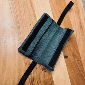 Può includere: Una custodia rettangolare in denim con due strisce di velcro nere e un cinturino nero. La custodia è appoggiata su una superficie di legno. La custodia sembra essere un supporto per il polso o la caviglia.