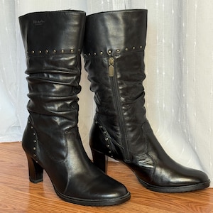 Peut inclure: Paire de bottes en cuir noir mi-mollet. Les bottes présentent un design froncé, une fermeture éclair et de petits clous dorés sur le dessus. Elles ont un talon bloc et un bout arrondi. Hauteur d'environ 30 cm.