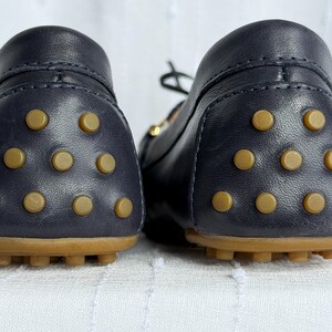 May include: A pair of navy blue leather driving shoes. The soles are brown with tan rubber studs. The uppers are smooth leather with a rounded toe and a small gold-colored accent.
