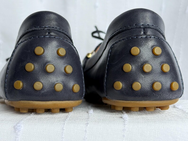 May include: A pair of navy blue leather driving shoes. The soles are brown with tan rubber studs. The uppers are smooth leather with a rounded toe and a small gold-colored accent.