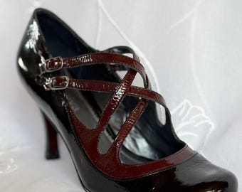May include: Black and burgundy Mary Jane style high-heeled shoe with a glossy finish. The shoe features a pointed toe, a crisscross strap design, and a small buckle closure. The heel is a matching burgundy color.