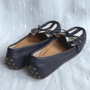May include: A pair of navy blue leather loafers with a white and black striped accent on the top. The shoes have a gold-colored bow detail and small gold studs on the heel.