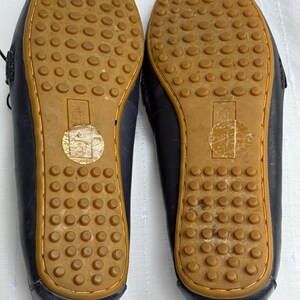 May include: A pair of navy blue leather driving shoes, soles up, with brown rubber soles featuring a textured pattern of small circles. The shoes have a stitched design around the edges.