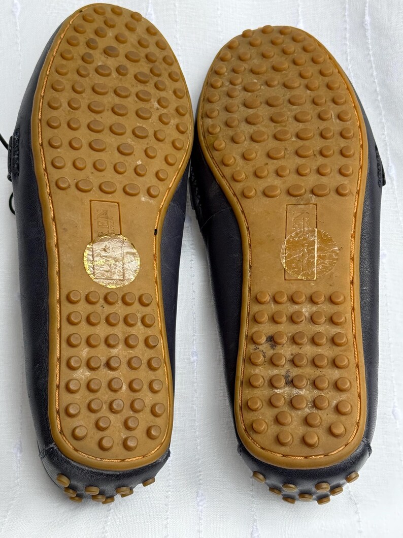 May include: A pair of navy blue leather driving shoes, soles up, with brown rubber soles featuring a textured pattern of small circles. The shoes have a stitched design around the edges.