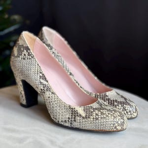 May include: A pair of vintage snakeskin-patterned high-heeled pumps. The shoes feature a pattern of grey, tan, and black. The heels are black, and the inner sole is pink. The shoes are displayed on a white surface.