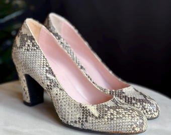 May include: A pair of vintage high-heeled pumps with a snakeskin pattern in shades of gray, tan, and black. The heels are a solid black, and the interior is a soft pink. The shoes are displayed on a white surface.
