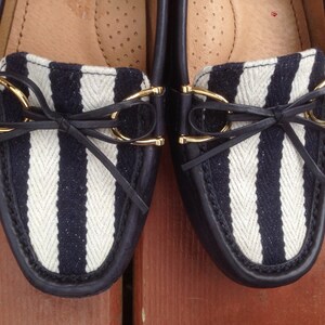 May include: Pair of navy blue and white striped loafers. The shoes have a leather trim, gold-colored accents, and a small bow detail. The interior is a light tan color.