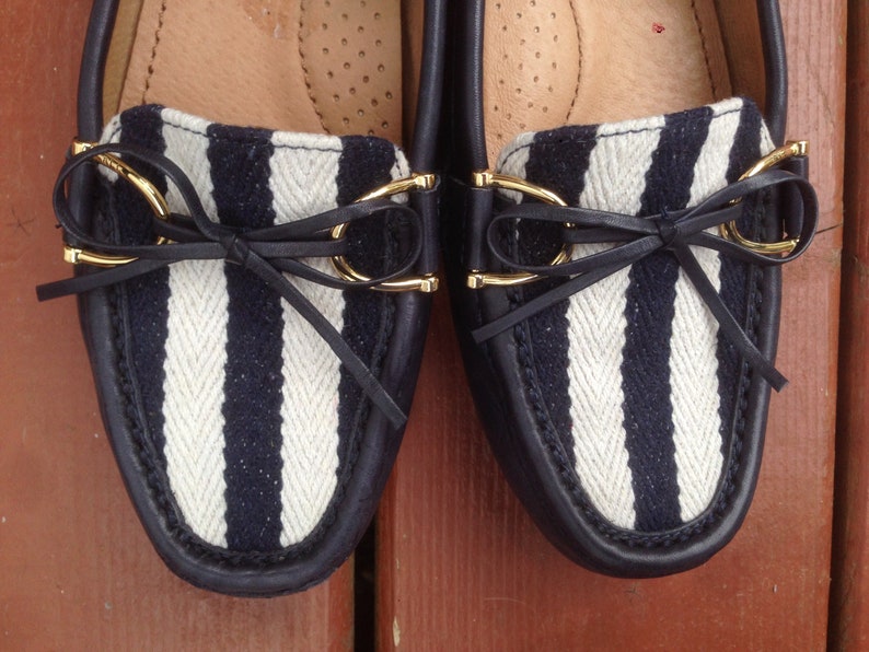 May include: Pair of navy blue and white striped loafers. The shoes have a leather trim, gold-colored accents, and a small bow detail. The interior is a light tan color.