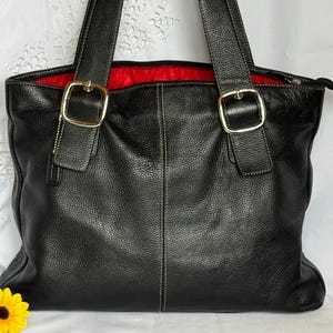 May include: Black leather tote bag with two top handles and gold-tone buckles. The bag has a red interior and white stitching details. A yellow flower is visible in the lower left corner.
