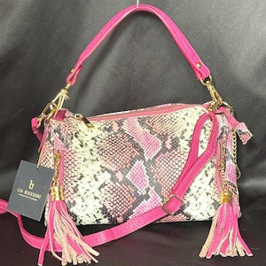 May include: A pink and white snakeskin-patterned handbag with a matching pink strap and gold hardware. The bag features a top handle, a detachable shoulder strap, and decorative tassels. A tag reads "LIA BIASSONI".
