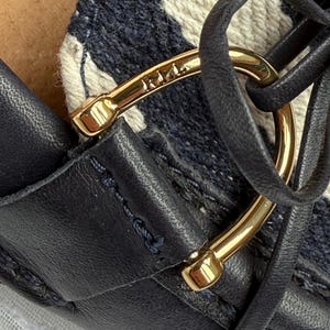 May include: Close-up of a navy blue leather shoe with a gold-colored metal accent. The shoe features a woven fabric pattern in white and navy blue. The gold accent has the word "Riri" engraved on it.