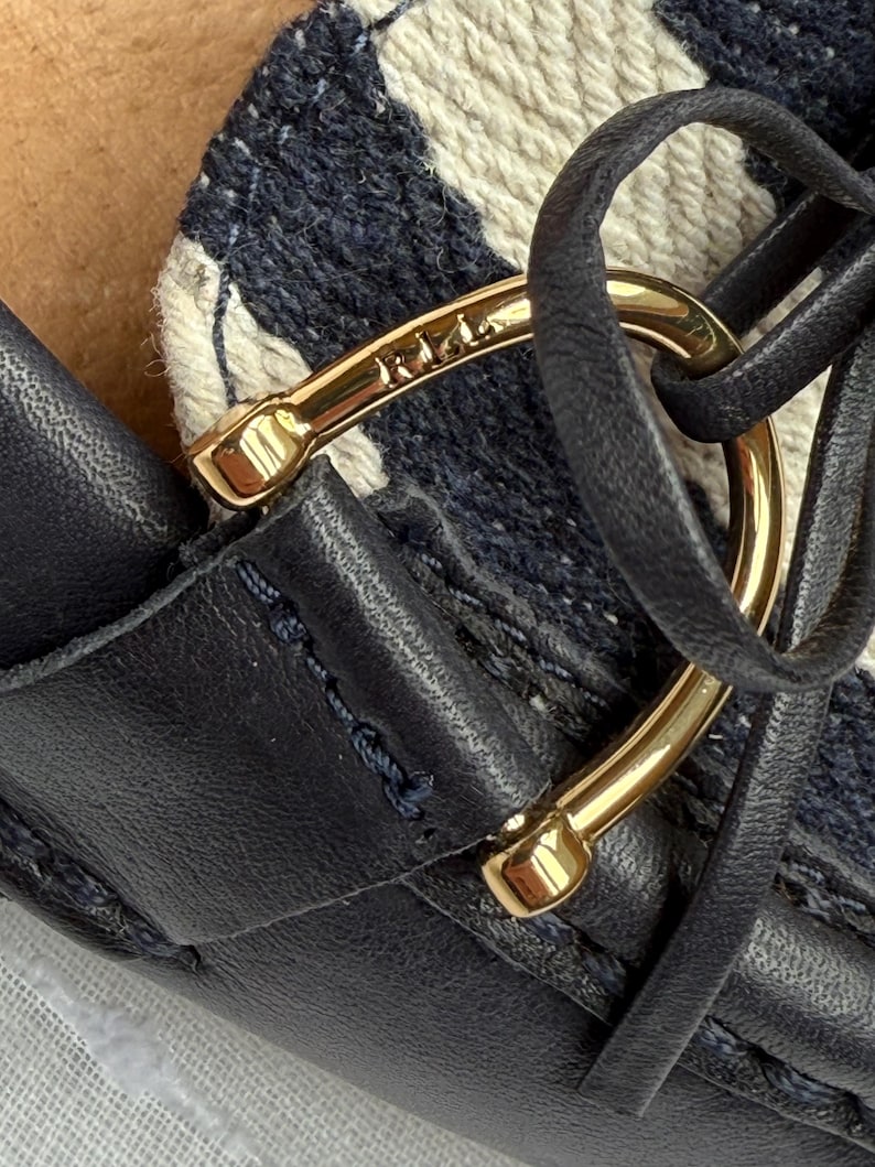 May include: Close-up of a navy blue leather shoe with a gold-colored metal accent. The shoe features a woven fabric pattern in white and navy blue. The gold accent has the word "Riri" engraved on it.