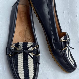 May include: A pair of navy blue leather loafers with a striped fabric detail on the vamp. The loafers have gold-colored accents and a bow. The soles are brown.