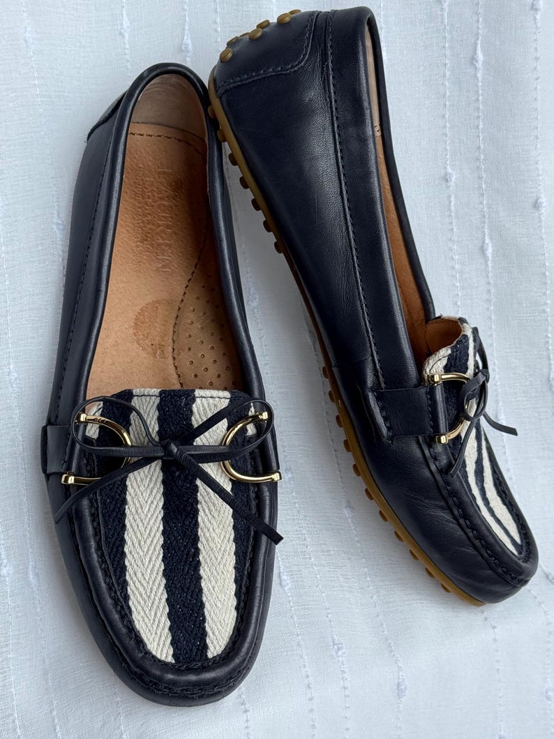 May include: A pair of navy blue leather loafers with a striped fabric detail on the vamp. The loafers have gold-colored accents and a bow. The soles are brown.