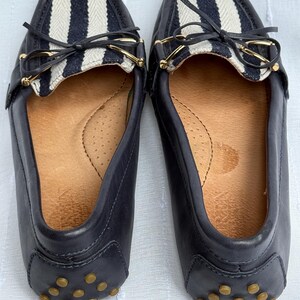 May include: A pair of navy blue leather loafers. The top of each shoe features a white and navy striped fabric panel with a small bow and gold-colored hardware. The soles have small, round, tan-colored rubber studs.
