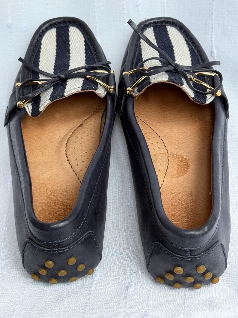 May include: A pair of navy blue leather loafers. The top of each shoe features a white and navy striped fabric panel with a small bow and gold-colored hardware. The soles have small, round, tan-colored rubber studs.