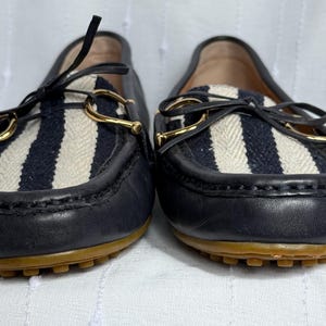 May include: Pair of navy blue leather loafers with a striped fabric panel in white and navy blue. The shoes have gold-colored metal accents and brown soles. The loafers are a classic style.