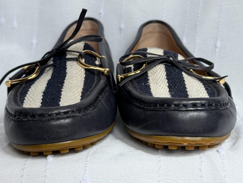 May include: Pair of navy blue leather loafers with a striped fabric panel in white and navy blue. The shoes have gold-colored metal accents and brown soles. The loafers are a classic style.