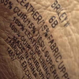 May include: Close-up of a textured, light brown leather surface with black text. The text includes "85% LEATHER/15% FABRIC UPPER", "BRILEY 6B", and "MADE IN CHINA". The image shows the material composition and origin of the product.