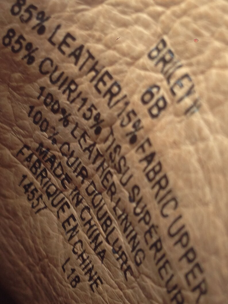 May include: Close-up of a textured, light brown leather surface with black text. The text includes "85% LEATHER/15% FABRIC UPPER", "BRILEY 6B", and "MADE IN CHINA". The image shows the material composition and origin of the product.