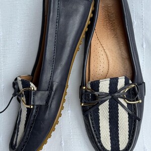 May include: A pair of navy blue leather loafers with a striped fabric detail in black and white. The shoes have a gold-colored buckle and a rubber sole. The loafers are designed for comfort and style.
