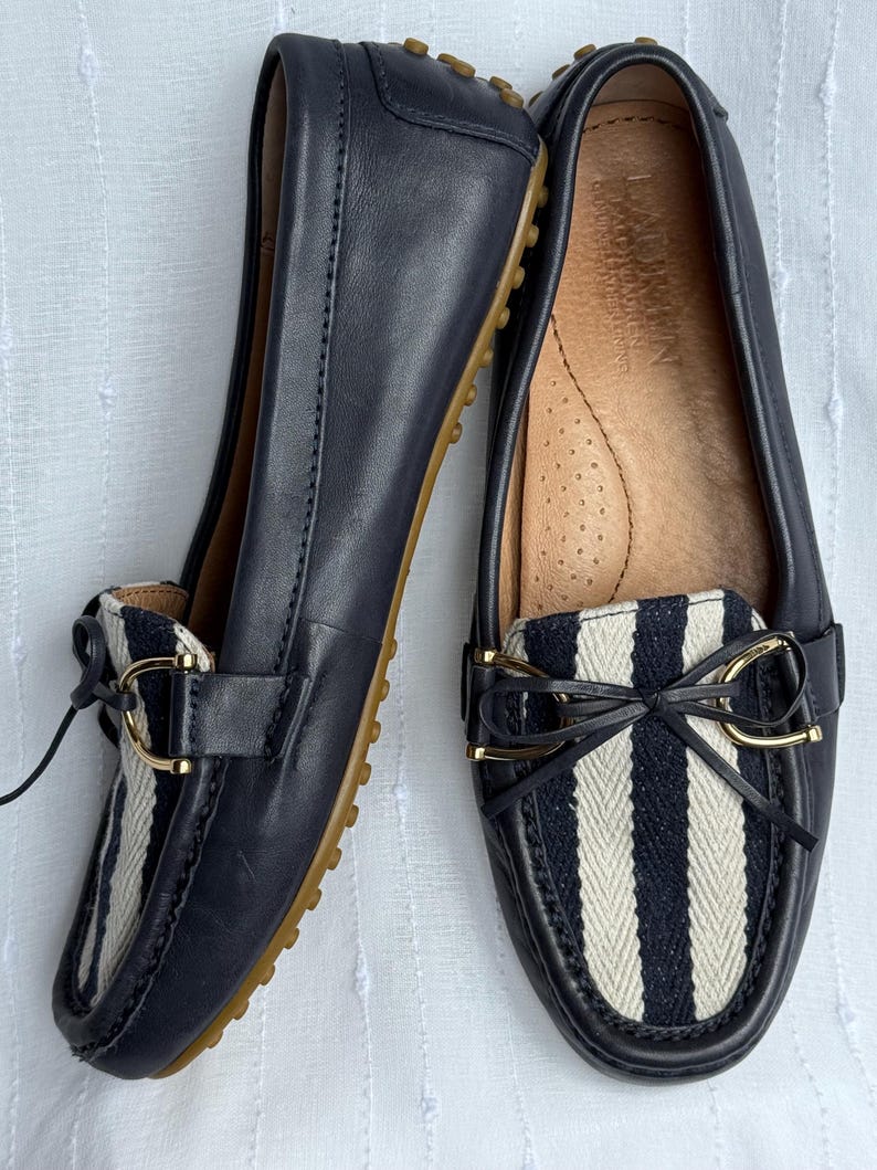 May include: A pair of navy blue leather loafers with a striped fabric detail in black and white. The shoes have a gold-colored buckle and a rubber sole. The loafers are designed for comfort and style.