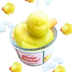 Puede incluir: Una vela amarilla con forma de pato en un vaso transparente, rodeada de pequeños derretidos de cera con forma de pato y derretidos blancos con forma de nube. El pato tiene un pico naranja y detalles rojos. El vaso tiene la inscripción "Hello Ducky!"