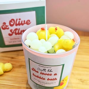 Puede incluir: Un tarro de velas con borde rosa lleno de ceras fundidas con forma de pato amarillo y nube blanca. La etiqueta del tarro dice "Smells like She loves a bubble bath". En el fondo, una caja con el texto "& Olive Almond".