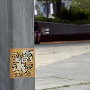 Può includere: Un adesivo su un palo di metallo presenta un unicorno dei cartoni animati con criniera e coda arcobaleno. L'unicorno tiene in mano un cartello con testo in tedesco. L'adesivo ha uno sfondo giallo con accenti floreali e stellati.