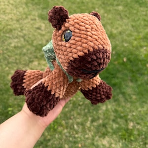 May include: A handmade crochet horse plushie in shades of brown and tan, with dark brown accents on the legs and ears. It has a green scarf and large, expressive eyes. The plush toy is soft and cuddly.