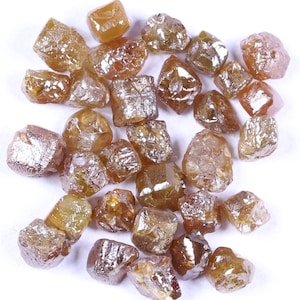 May include: A collection of rough, uncut diamonds in shades of brown and yellow. The gemstones have a natural, irregular shape and a sparkling, reflective surface. The diamonds are scattered on a white background.