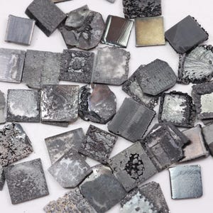 Op de afbeelding: Een verzameling kleine, onregelmatig gevormde metalen vierkanten en rechthoeken in verschillende tinten grijs en zilver. Sommige stukken hebben een getextureerd, ruw oppervlak, terwijl andere glad en reflecterend zijn. De stukken zijn verspreid op een wit oppervlak.