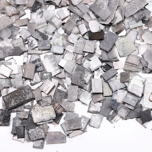 Puede incluir: Un conjunto de pequeños fragmentos de metal gris de formas irregulares, dispersos sobre un fondo blanco. Las piezas presentan una gama de tonos grises, algunos con texturas. La imagen sugiere un tema industrial o abstracto.