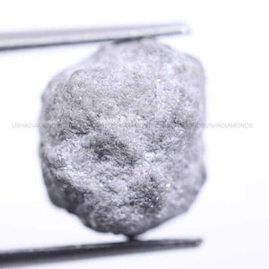 Puede incluir: Un diamante gris en bruto sin tallar sostenido por pinzas metálicas. La piedra presenta una superficie texturizada e irregular con un acabado mate y facetas naturales. El texto USHADIAMONDS es ligeramente visible en el centro.