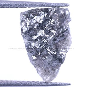 May include: A raw, uncut grey diamond with a multifaceted, crystalline surface texture. The stone exhibits a dark, metallic lustre and irregular, angular edges. The text USHADIAMONDS is visible across the centre of the stone in small print.