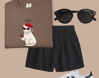 May include: A casual outfit featuring a brown t-shirt with a cat graphic, black shorts, black sunglasses, and white sneakers with black stripes. The t-shirt has the text "J'adore."