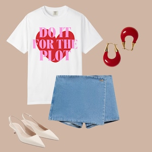 Puede incluir: Una camiseta blanca con el texto "DO IT FOR THE PLOT" en rosa y rojo, combinada con una falda vaquera, tacones blancos y pendientes rojos. Los pendientes tienen un acento dorado.
